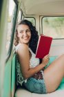 Smiling young woman sitting inside caravan and reading with book — Stock Photo