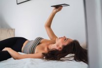 Jeune femme brune en haut à rayures et collants noirs couchés sur le lit dans la chambre et à l'aide d'un smartphone — Photo de stock