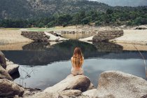 Visão traseira da jovem mulher com cabelos longos em terno de natação sentado em pedra grande e olhando ainda lago no fundo em árvores verdes e montanhas — Fotografia de Stock