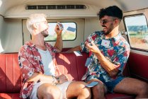 Два веселых молодых парня смеются и бросают игральные карты, сидя на пассажирском сидении ретро-фургона во время поездки на природу — стоковое фото
