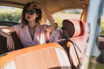 Hermosa mujer confiada en rosa y gafas de sol sentado en el asiento trasero en coche retro mirando hacia otro lado a la luz del sol - foto de stock