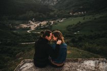 Linda pareja abrazando y endureciendo la frente mientras se sienta en la ladera rocosa en el fondo del hermoso valle y las montañas - foto de stock