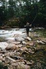 Pescador segurando vara giratória e em pé sobre rochas no fluxo do rio — Fotografia de Stock