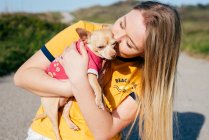 Happy casual girl with long blond hair holding and kissing small dog Chihuahua standing on road in sunshine. — Stock Photo