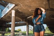Young African American woman in denim shirt and shorts standing under bridge with hand in hair and looking at camera — Stock Photo