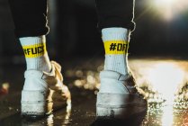 Piernas de persona irreconocible en zapatillas con hashtags Fuck Off en calcetines por la noche - foto de stock