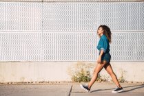 Молодая афроамериканка с темными волосами в джинсовой одежде и кроссовках, идущая по улице возле стены в солнечный день — стоковое фото
