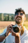 Handsome young guy in sunglasses cheerfully smiling and holding retro photo camera while standing on blurred background of amazing countryside — Stock Photo