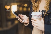 Unrecognizable woman holding smartphone and paper cup on blurred street — Stock Photo