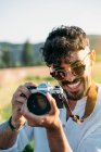 Bonito cara jovem em óculos de sol alegremente sorrindo e segurando câmera de foto retro enquanto está de pé no fundo borrado do campo incrível — Fotografia de Stock