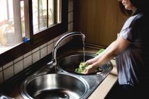 Vue de côté de la récolte de la femme laver la salade verte dans un évier en acier debout dans la cuisine — Photo de stock