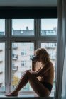 Mujer joven rubia desnuda bebiendo café mientras está sentada en el alféizar de la ventana por la mañana - foto de stock