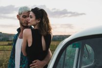 Embracing man and woman in elegant dress standing near car on background of landscape — Stock Photo