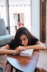 Jovem mulher pensativa sentada à mesa pequena de madeira no quarto e escrevendo no diário — Fotografia de Stock
