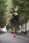 Рыжая балерина с черной пачкой и красными балетными кончиками танцует на улице с деревьями на заднем плане . — стоковое фото