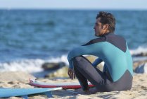 Rückansicht eines Mannes im Neoprenanzug, der mit Surfbrett am Strand sitzt und auf das Meer blickt. — Stockfoto
