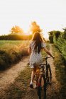 Back view of pretty woman carrying bicycle and walking on rural road in backlit — Stock Photo
