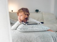 Вдумчивый мальчик лежит с цифровой табличкой на диване и смотрит в сторону — стоковое фото