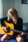 Bionda giovane donna che suona la chitarra sul letto a casa — Foto stock