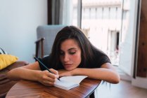 Молода жінка вдумливі, сидячи на дерев'яні невеликий столик в кімнаті та запис у щоденнику — стокове фото