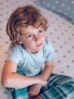 Charming boy in pajamas sitting on comfortable bed and looking away — Stock Photo