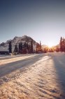 Яркое оранжевое солнце, сияющее над полем с домами и деревьями, покрытыми снегом на заднем плане с живописными горами и ясным небом — стоковое фото