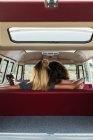 Задний вид двух женщин, обнимающих друг друга, сидя на заднем сиденье ретро-фургона в природе — стоковое фото