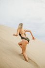 Jovem loira em roupa de banho preta correndo duna de areia enquanto descansa na praia — Fotografia de Stock
