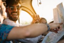 Dois jovens rindo e segurando mapa enquanto sentados dentro do carro retro durante uma viagem agradável — Fotografia de Stock
