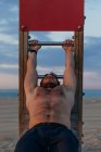 Shirtless muscular guy doing abdominal crunches on wooden slide on beach — Stock Photo