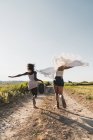 Веселі стильні багатоетнічні жінки з валізою і шарфом з ентузіазмом бігають по дорозі в літній зеленій сільській місцевості — стокове фото