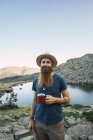 Бородатий молодий чоловік стоїть біля озера в горах з чашки і дивлячись на камеру — стокове фото
