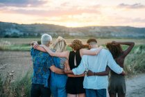 Back view of young men and women embracing each other and admiring beautiful sunset while spending time in nature together — Stock Photo