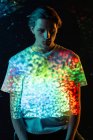 Modèle masculin androgyne regardant des taches lumineuses de lumière rouge et bleue sur le T-shirt tout en se tenant debout sur fond noir — Photo de stock