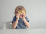 Adorabile ragazzo fare faccia divertente con due fette di limone invece di occhi mentre seduto su sfondo bianco — Foto stock