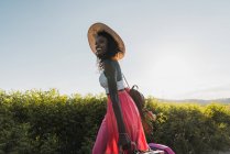 Trendy African-American woman in hat carrying suitcase and walking on rural road in summertime — Stock Photo