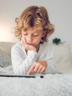 Скучный мальчик тыкает цифровым экраном планшета, лежа на удобном диване — стоковое фото