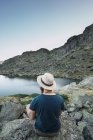 Молодий чоловік в капелюсі сидить на каменях біля озера і дивиться на погляд — стокове фото