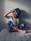Очаровательный мальчик в пижаме с книгой на голове и с удовольствием читает, сидя на удобной кровати — стоковое фото