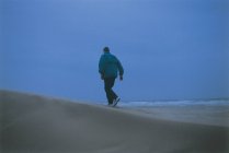 Vue arrière de l'homme en veste chaude et pantalon marchant sur la plage de sable près de la mer sur fond de ciel bleu sans nuages — Photo de stock