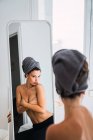 Jeune femme seins nus debout devant le miroir avec une serviette sur la tête — Photo de stock