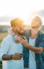 Deux hommes souriants étreignant et naviguant smartphones tout en se tenant dans la belle campagne ensemble — Photo de stock