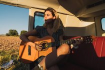 Mulher tocando guitarra acústica enquanto sentada dentro de van retro durante a viagem — Fotografia de Stock