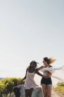 Веселые стильные мультиэтнические женщины с чемоданом и шарфом, с волнением бегущие по дороге в летней зеленой сельской местности — стоковое фото