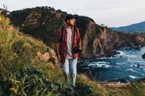 Красивый молодой парень с фотокамерой смотрит в сторону, стоя на скале возле воды во время поездки на природу — стоковое фото