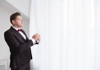 Вид сбоку на молодого красивого мужчину, стоящего в комнате в черном костюме и смотрящего через окно за белым занавесом — стоковое фото