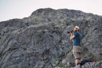 Jovem homem de chapéu em pé perto de penhasco rochoso e tirar foto com câmera fotográfica — Fotografia de Stock