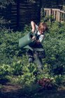 Garçon avec les cheveux bouclés dans les plantes d'arrosage de tablier dans le jardin — Photo de stock