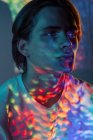 Young androgynous guy illuminated with colorful sports of bright light looking away while standing on black background — Stock Photo