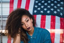 Jovem mulher pensativa sentada contra a bandeira da América — Fotografia de Stock